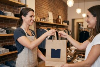 Commerçante tendant un sac papier personnalisé à une cliente dans une boutique au décor chaleureux