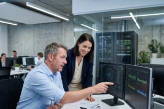 Responsable informatique interne discutant avec une collègue devant des écrans affichant des schémas réseau dans un bureau de PME moderne